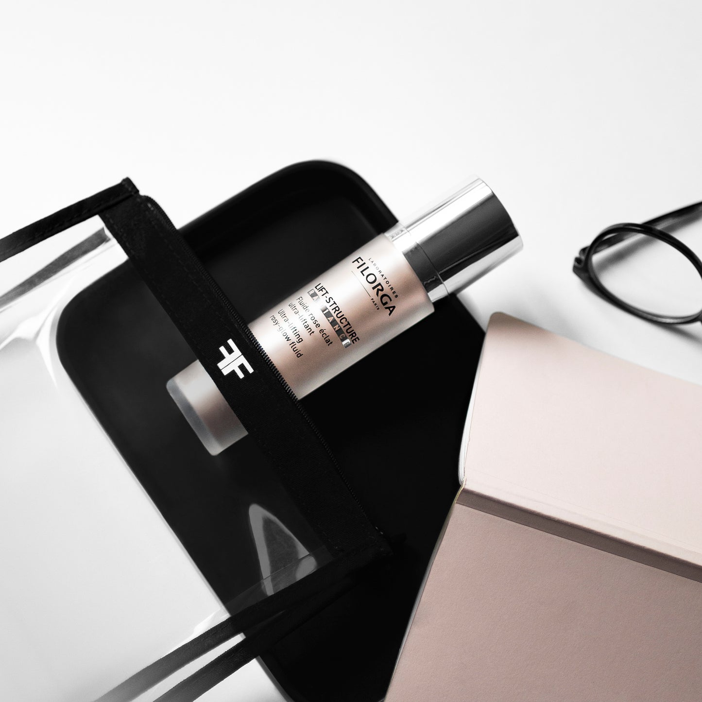 FILORGA LIFT-STRUCTURE RADIANCE bottle diagonally placed on a table near glasses and a FF FILORGA clear pouch