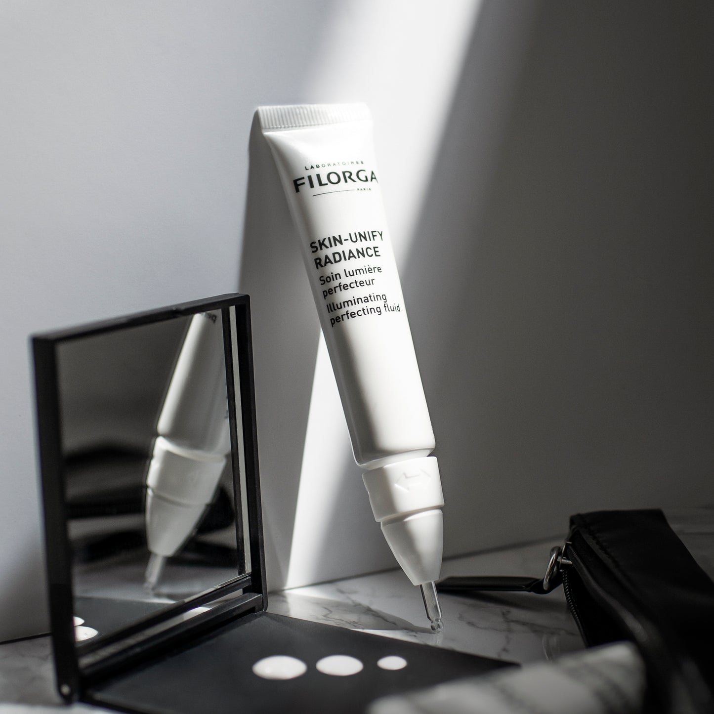 Open SKIN-UNIFY RADIANCE tube leaning against a wall with dots of serum on compact mirror