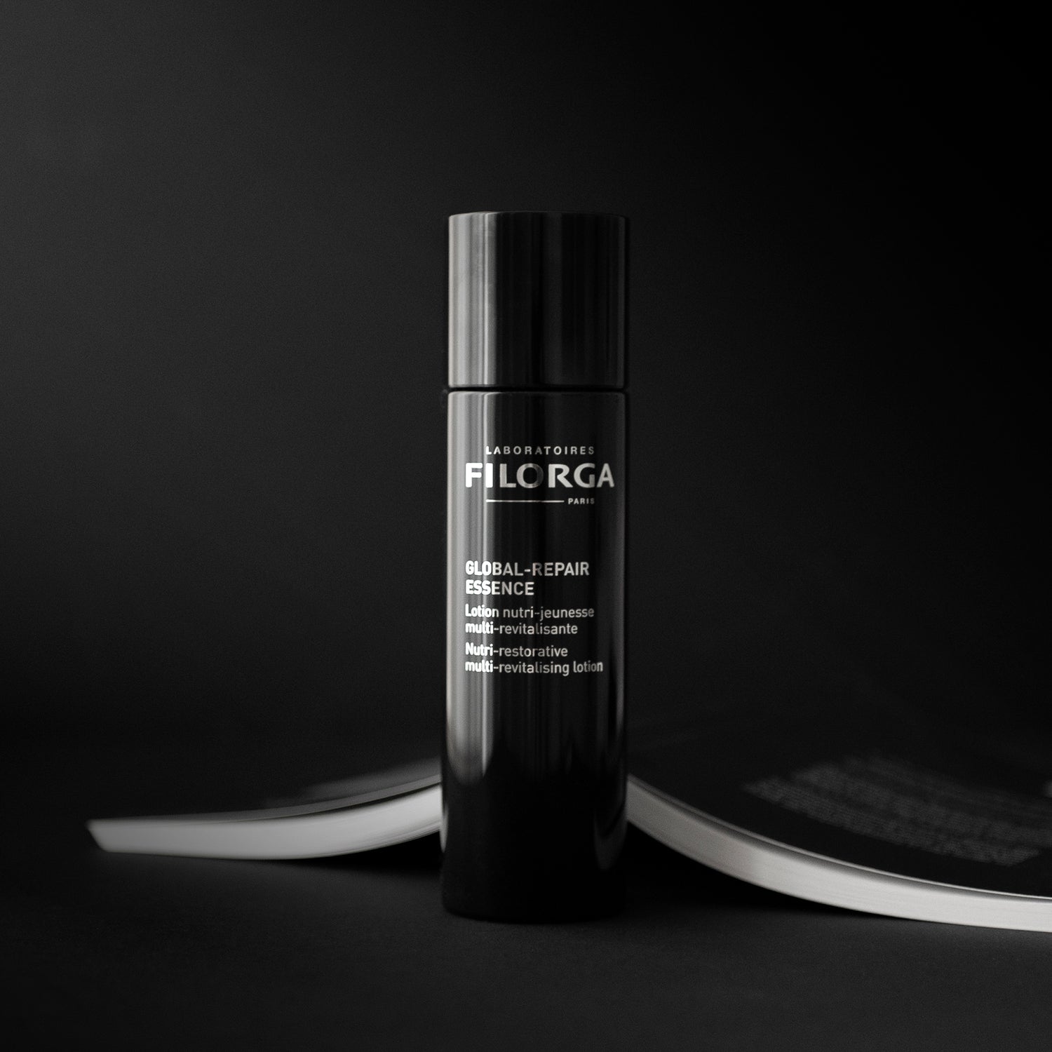 FILORGA GLOBAL-REPAIR ESSENCE closed black bottle next to a face down open black book