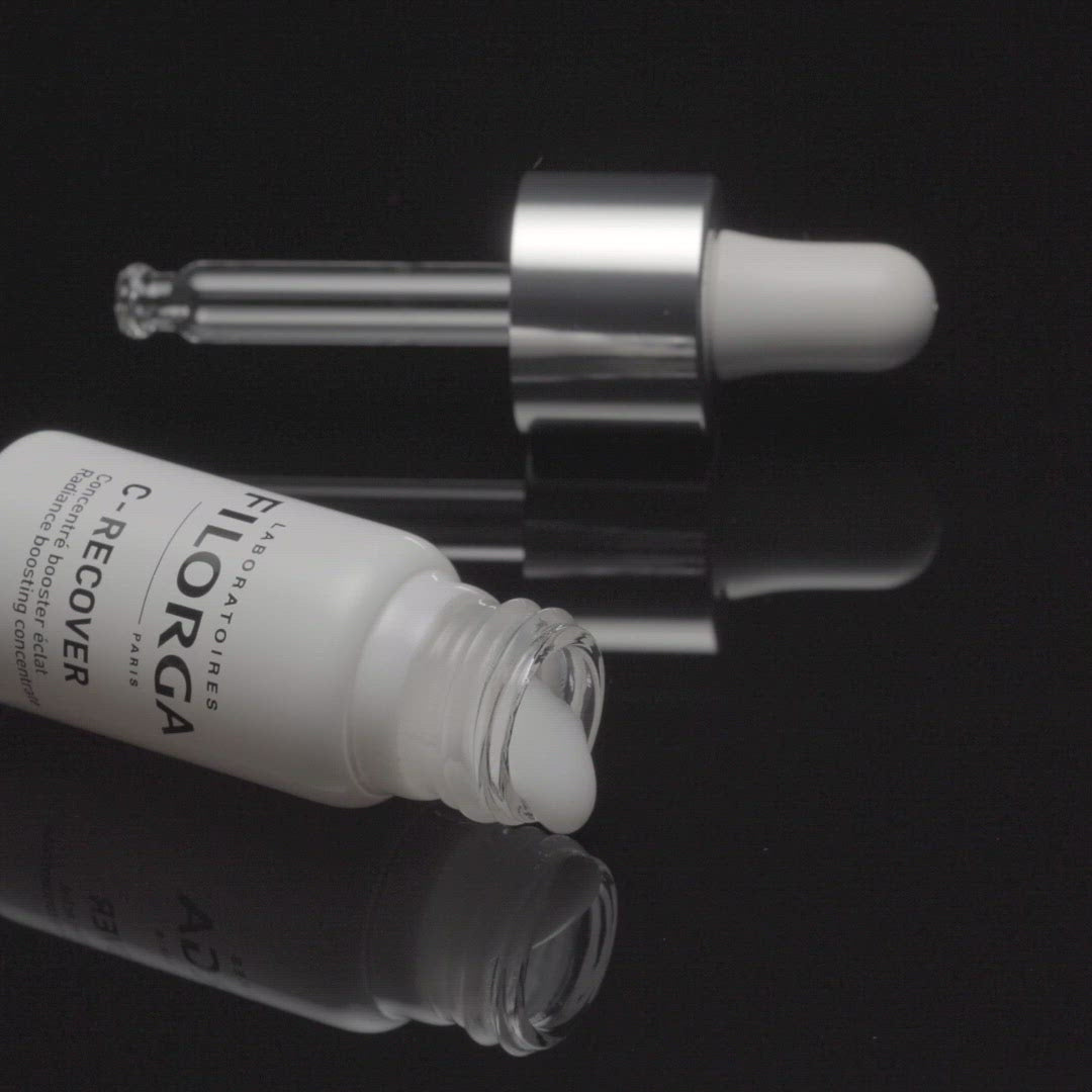 FILORGA C-RECOVER dropper and white bottle lying ona black table with product spilling out